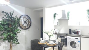 Eine moderne Küche mit Mikrowelle, Backofen und Waschmaschine. Ein Esstisch mit Stühlen und eine Uhr an der Wand sind vorhanden.