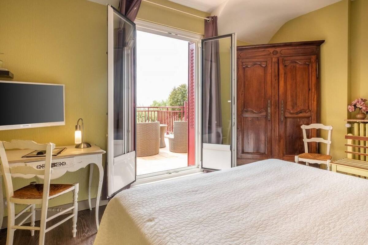 A bedroom with a bed, a desk, a chair, a wardrobe, and a balcony with wicker furniture.