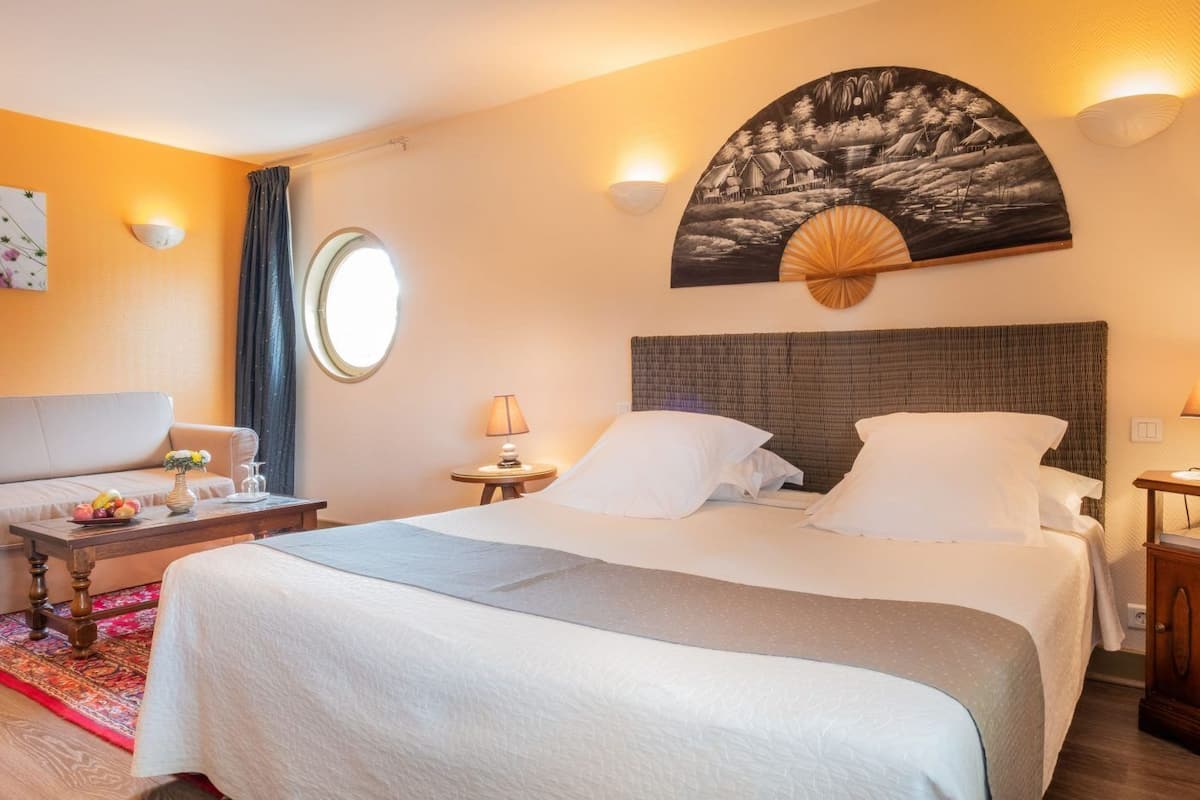 A hotel room with a bed, a sofa, a round window, and a wall-mounted artwork.