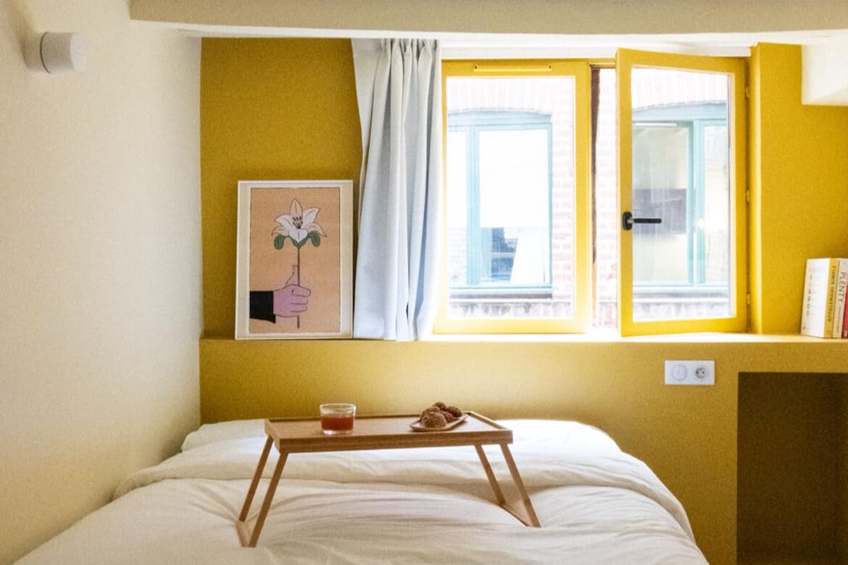 A room with a bed, a window with curtains, a small table with a tray, and a picture on the wall.