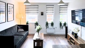 Una sala de estar moderna con un sofá negro, una mesa de centro de madera y un televisor montado en la pared. Hay tres ventanas con persianas, un radiador y una planta en maceta.