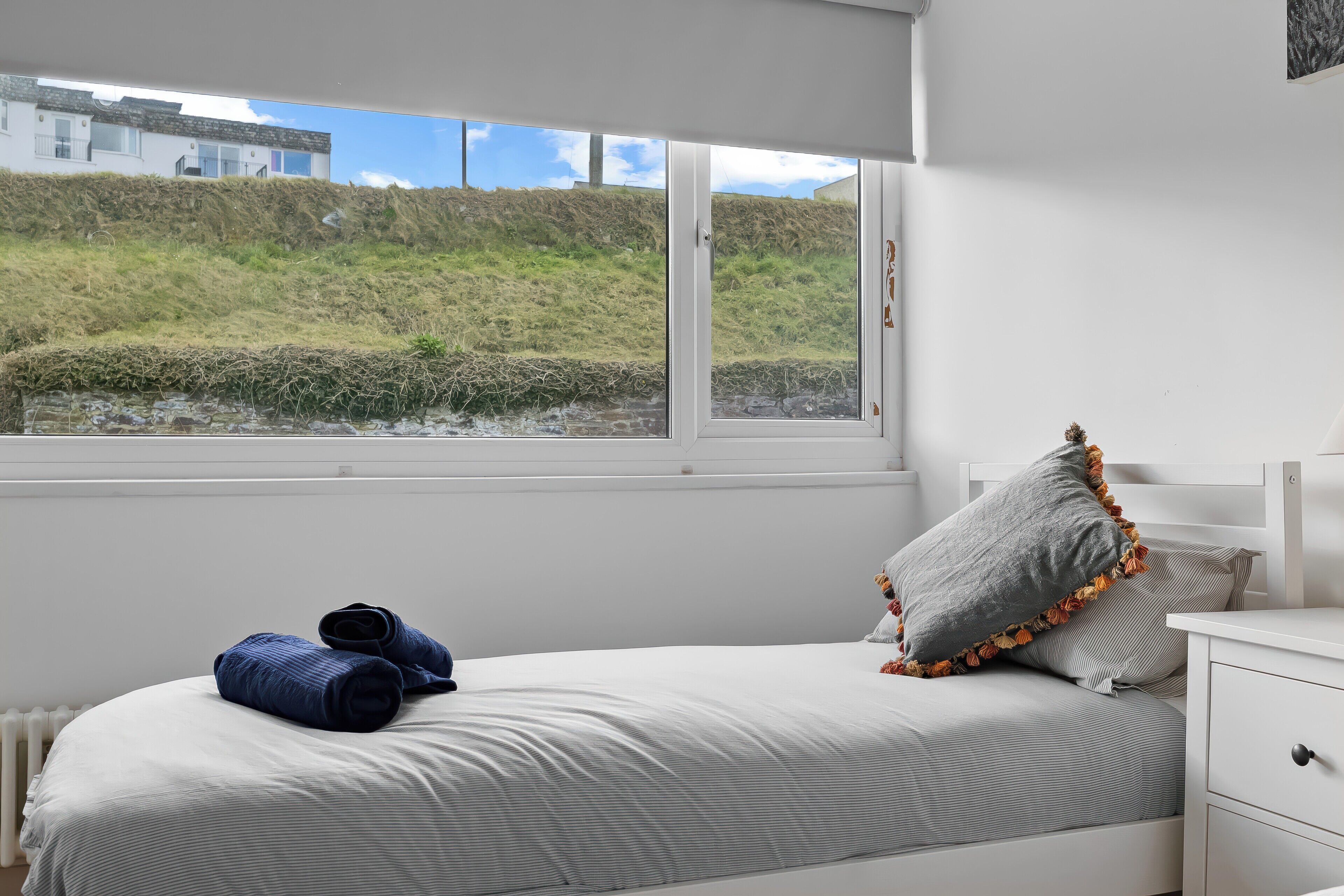 Un dormitorio con cama, almohadas, una mesita de noche y una ventana con vistas a una colina cubierta de hierba.