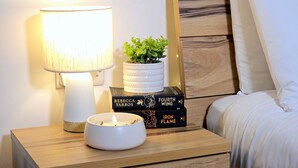 Ein Nachttisch mit einer weißen Lampe, einem kleinen Topfpflanzen und einer Schale. Auf dem Tisch liegen Bücher gestapelt, und im Hintergrund ist ein Bett mit weißen Laken zu sehen.