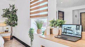 A modern room with a laptop on a wooden desk, a large potted plant, and a window with blinds.