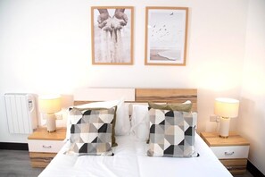 Een net opgemaakt bed met geometrische kussens, een nachtkastje met een lamp, en twee ingelijste foto's aan de muur.