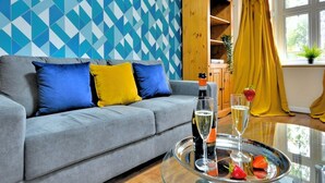 Una sala de estar con un sofá gris, cojines azules y amarillos, una mesa de vidrio con una botella y vasos, y una pared con un estampado geométrico.