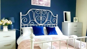 Un dormitorio con estructura de cama metálica blanca, colcha rosa y almohadas azules. Hay una mesita de noche blanca con un jarrón de flores y una lámpara. Encima de la cama, cuelga un cuadro enmarcado.