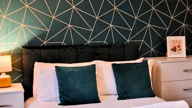 Una cama bien hecha con almohadas blancas y verde oscuro, una manta color beige y toallas blancas enrolladas, con una pared de fondo con diseño geométrico.
