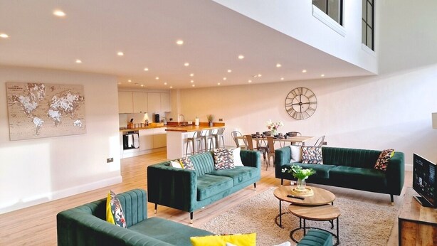 A spacious living area with green sofas, a coffee table, and a TV. There is a kitchen in the background with a dining area.