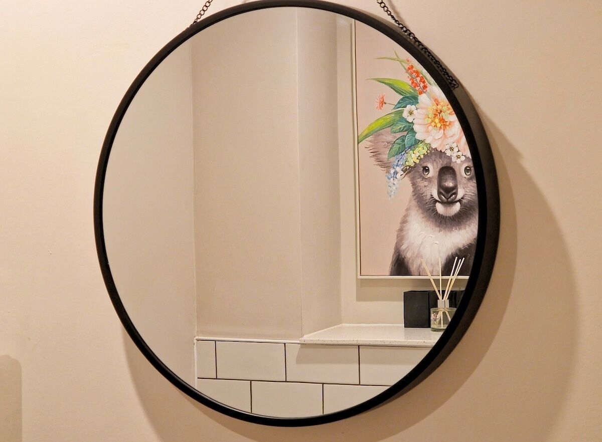 Un miroir rond doté d’un cadre noir, dans lequel se reflète une œuvre d’art murale représentant un koala portant une couronne de fleurs.