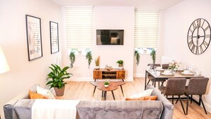 A modern living room with a sofa, coffee table, and dining area. There are two potted plants, a TV mounted on the wall, and a large clock.