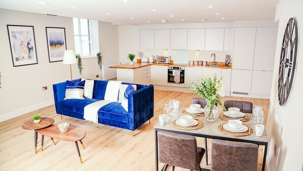 A modern living room with a blue sofa, a dining table set with white plates and glasses, and a kitchen area with white cabinets.