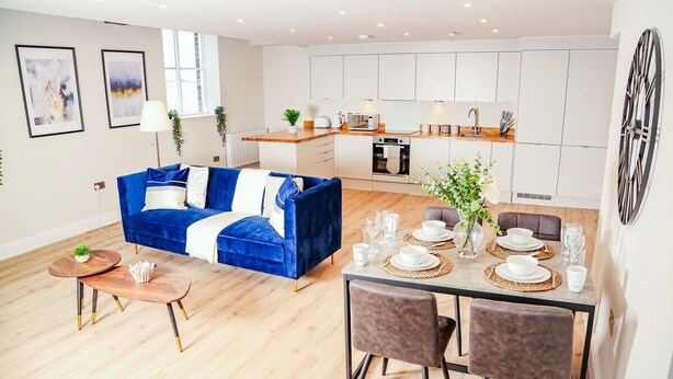 A modern living room with a blue sofa, a dining table set for four, and a kitchen in the background. - Shrewsbury 1 Bedroom Flat By Bevolve (Shrewsbury)