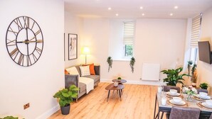 A modern living room with a sofa, a coffee table, and a dining area set for four. There is a large wall clock, a window with blinds, and several potted plants. - Shrewsbury 1 Bedroom Flat By Bevolve (Shrewsbury)