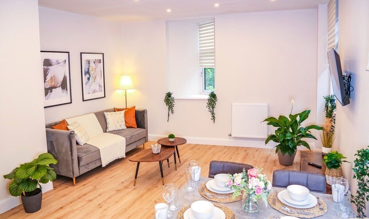 A modern living room with a grey sofa, a coffee table, and a dining area set with plates and glasses. There are two framed pictures on the wall, a lamp, and a TV mounted on the wall.