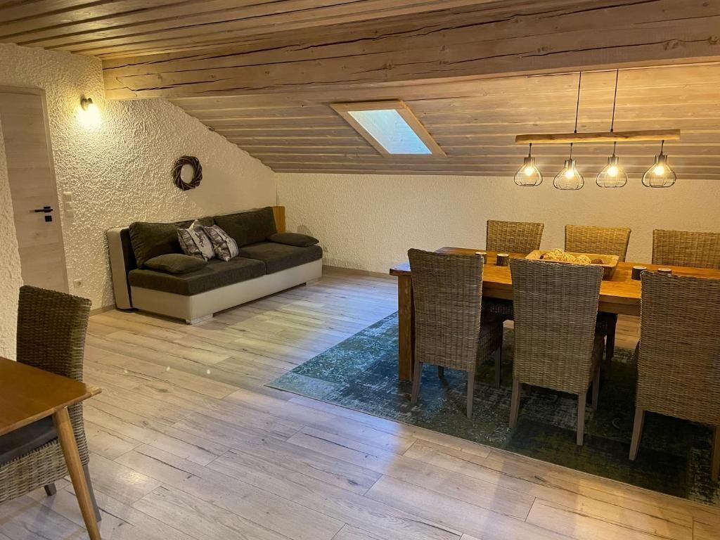 A cozy room with a wooden ceiling, a sofa with cushions, a dining table with wicker chairs, and a small rug.
