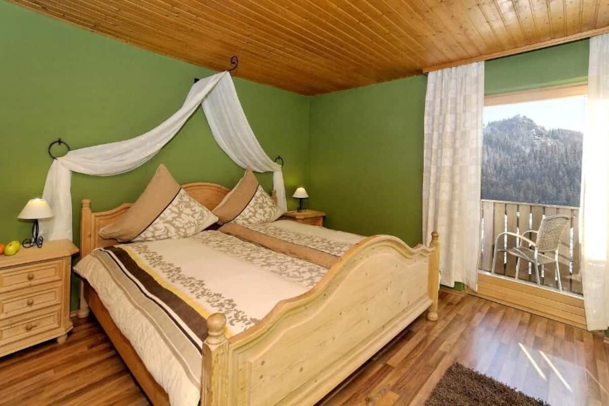 Een slaapkamer met een houten bed, groene wanden, een balkon met een stoel en uitzicht op een berg.