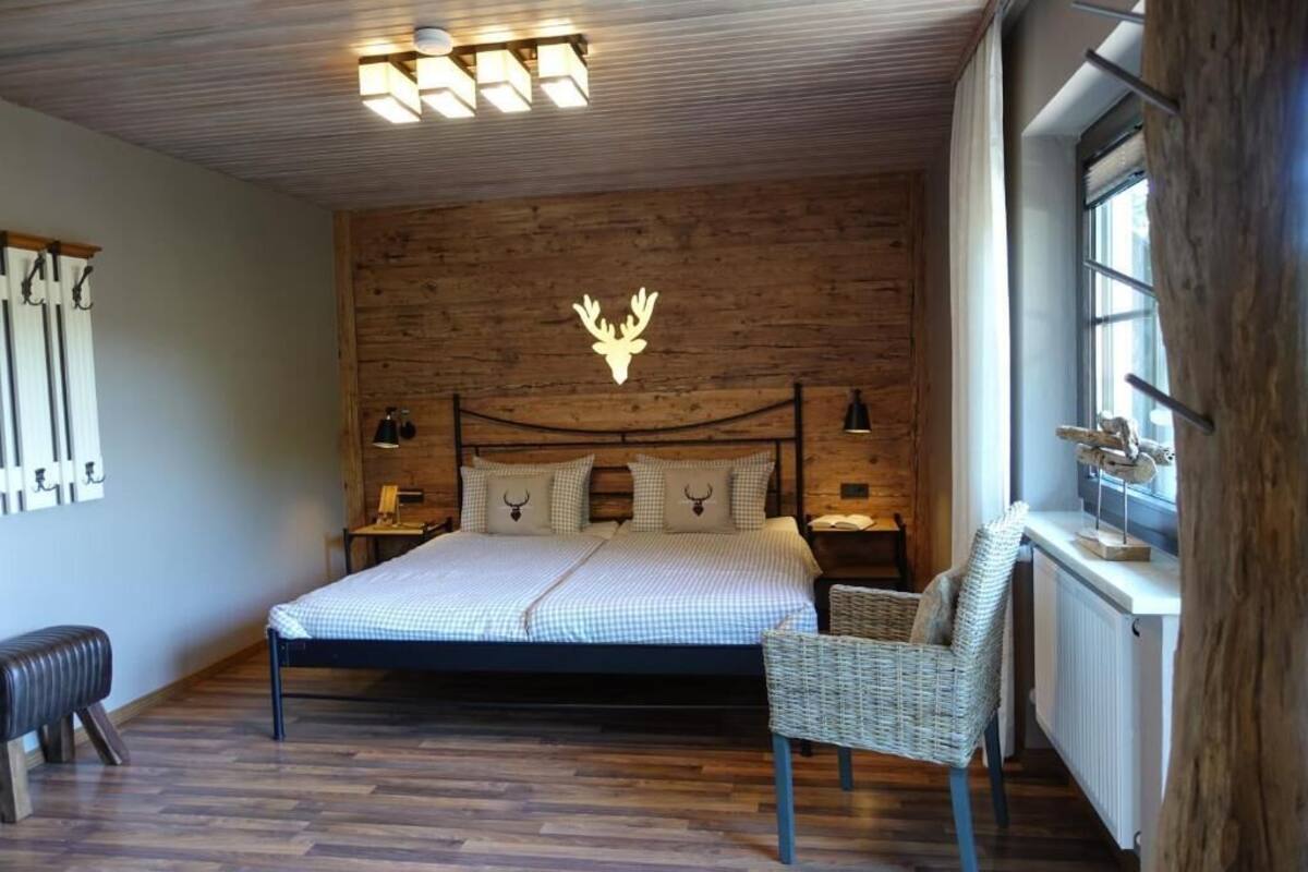 Een slaapkamer met een houten hoofdbord, een bed met geruit beddengoed, een rieten stoel en een nachtkastje met een lamp.