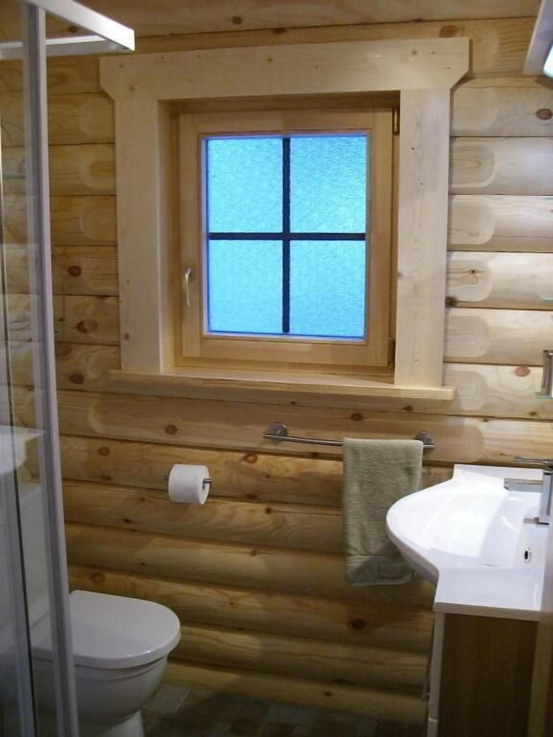 Une salle de bain rustique, aménagée dans une cabane en bois, avec une fenêtre, des toilettes, un lavabo et une serviette accrochée au mur.