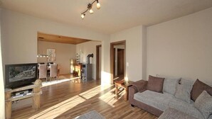 A spacious living room with a wooden floor, a grey sofa, a television on a wooden stand, and a kitchen area in the background. - Montara Suites Bodenmais (Bodenmais)