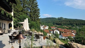 An outdoor patio with tables and chairs, surrounded by greenery and overlooking a village. - Montara Suites Bodenmais (Bodenmais)