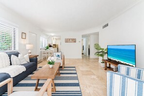 Una sala de estar con sofá, mesa de centro y televisor, una cocina al fondo y un cartel con la palabra 'BEACH'.