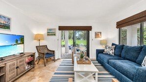 Una sala de estar con televisor, sofá, sillón y mesa de centro. Se ve un jardín a través de un ventanal.