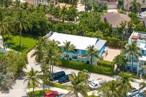 A residential area with a large house featuring a blue and white color scheme, surrounded by palm trees and other greenery.