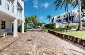 A paved walkway leading to a building with a white facade and balconies, surrounded by greenery and palm trees.