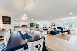 A spacious living area with a dark wooden dining table, white chairs, a blue sofa set, and a kitchen in the background. - The Sandstone at Juno Beach | 11 | Rooftop Deck! (Juno Beach)