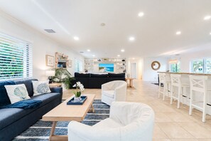 Una sala de estar moderna con sofá, mesa de centro y sillas, un área de cocina con televisor, y una barra de comedor con sillas.