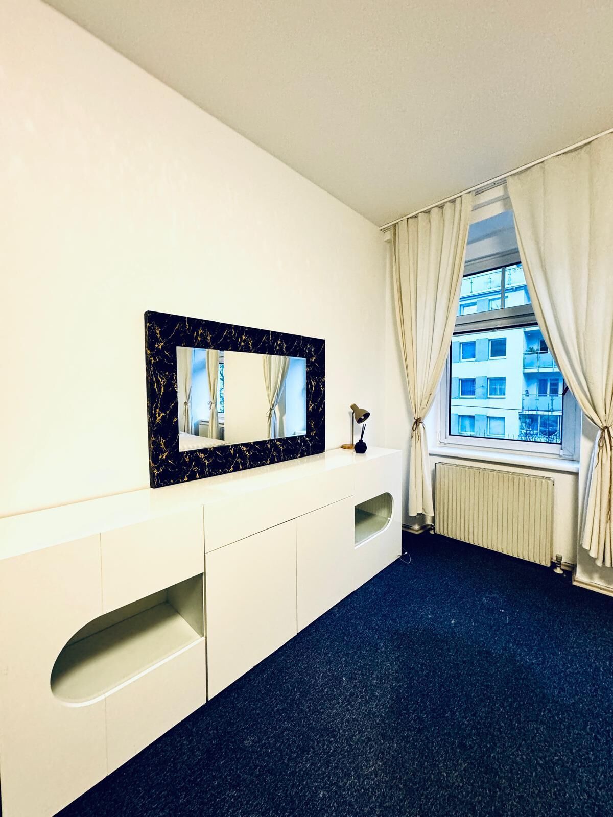 A hotel room with a large mirror, a white dresser, a radiator, and a window with curtains.