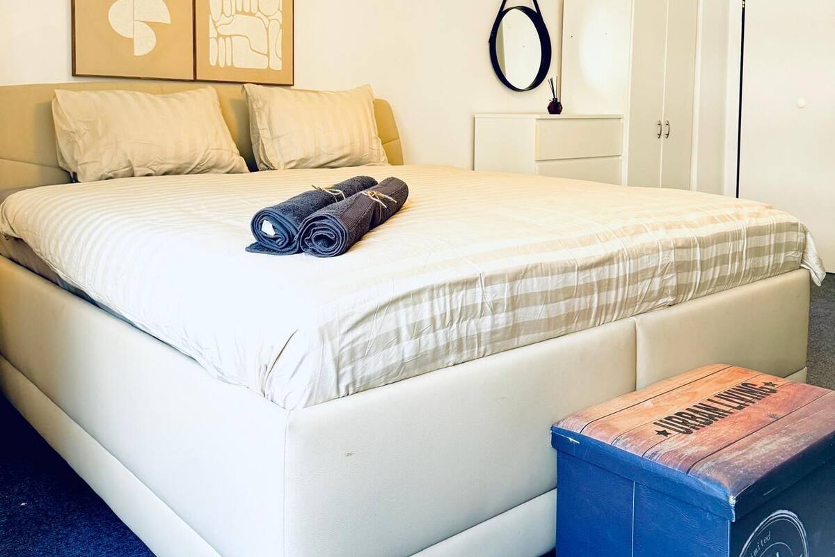 A neatly arranged bedroom with a bed, pillows, a rolled-up towel, a bedside table, a mirror, and a small box.