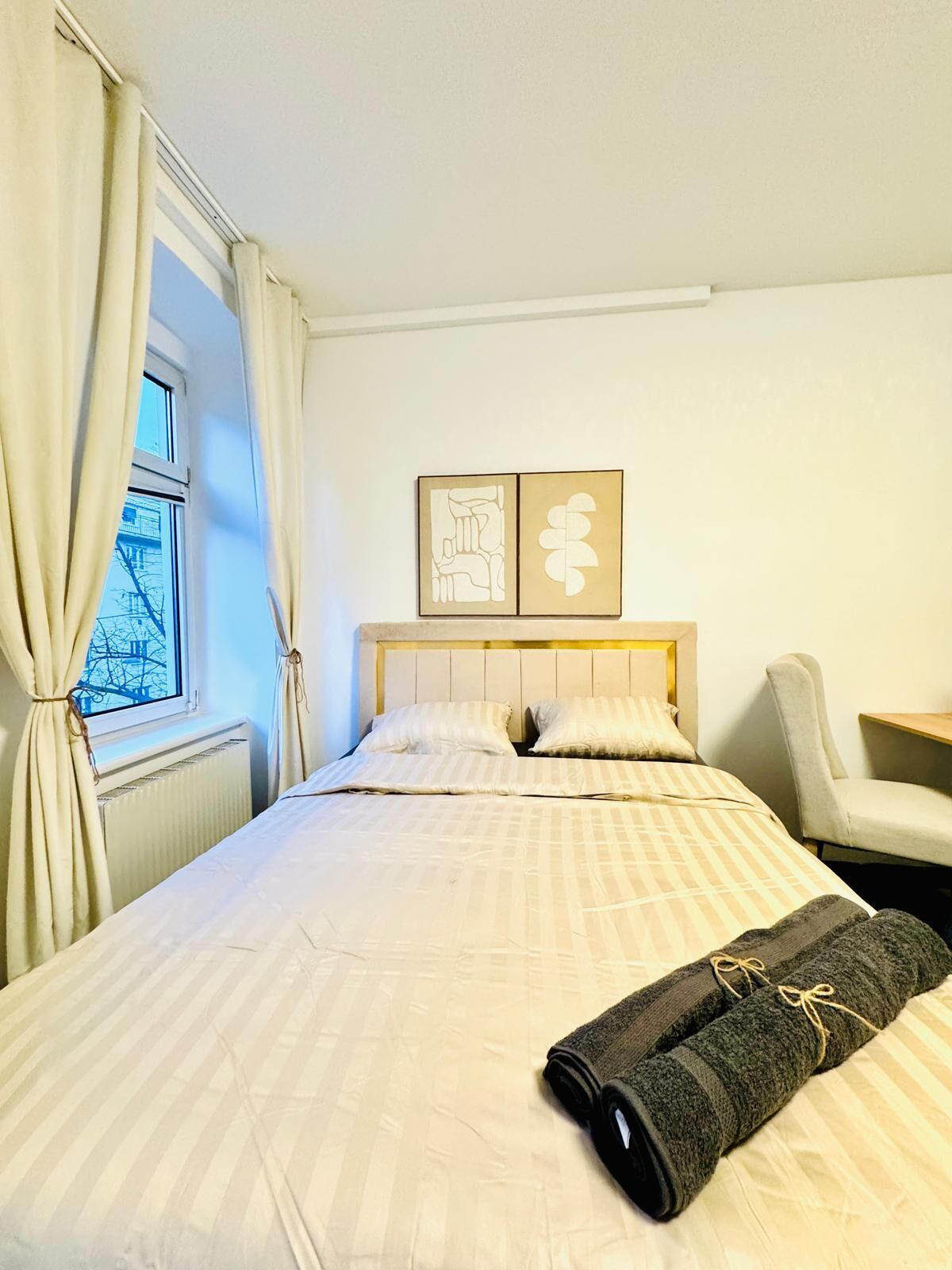 A hotel room with a bed, a chair, a radiator, and a window with curtains.