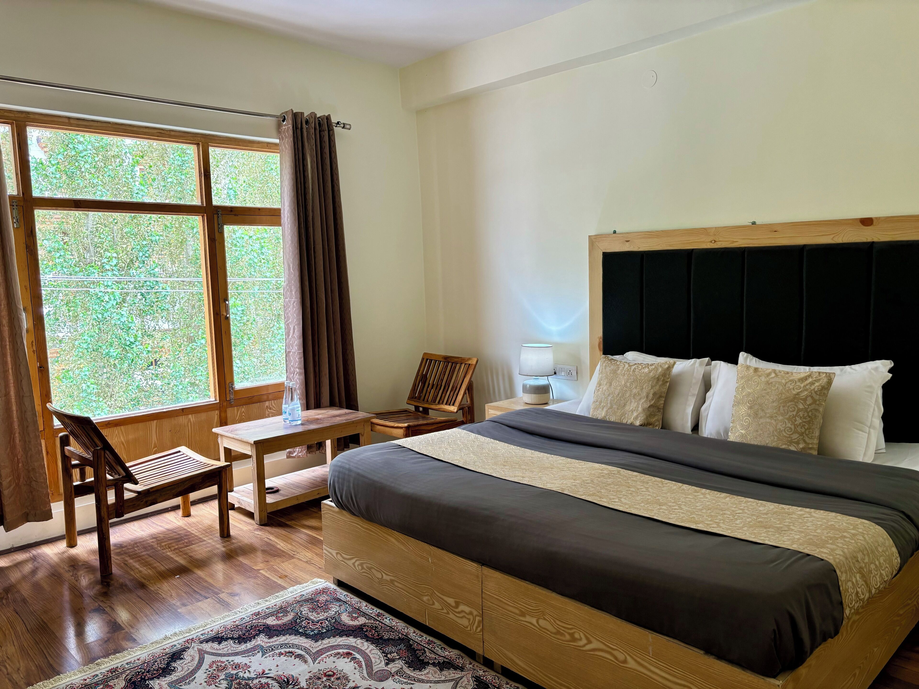 Un dormitorio con una cama grande, cabecera de madera, mesita de noche con lámpara, una silla, una pequeña mesa con una botella y una ventana con cortinas.