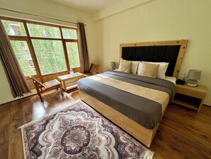 Une chambre à coucher avec un grand lit, une tête de lit en bois, une table de chevet avec une lampe, une petite table avec des chaises, et une vue sur la verdure par la fenêtre.