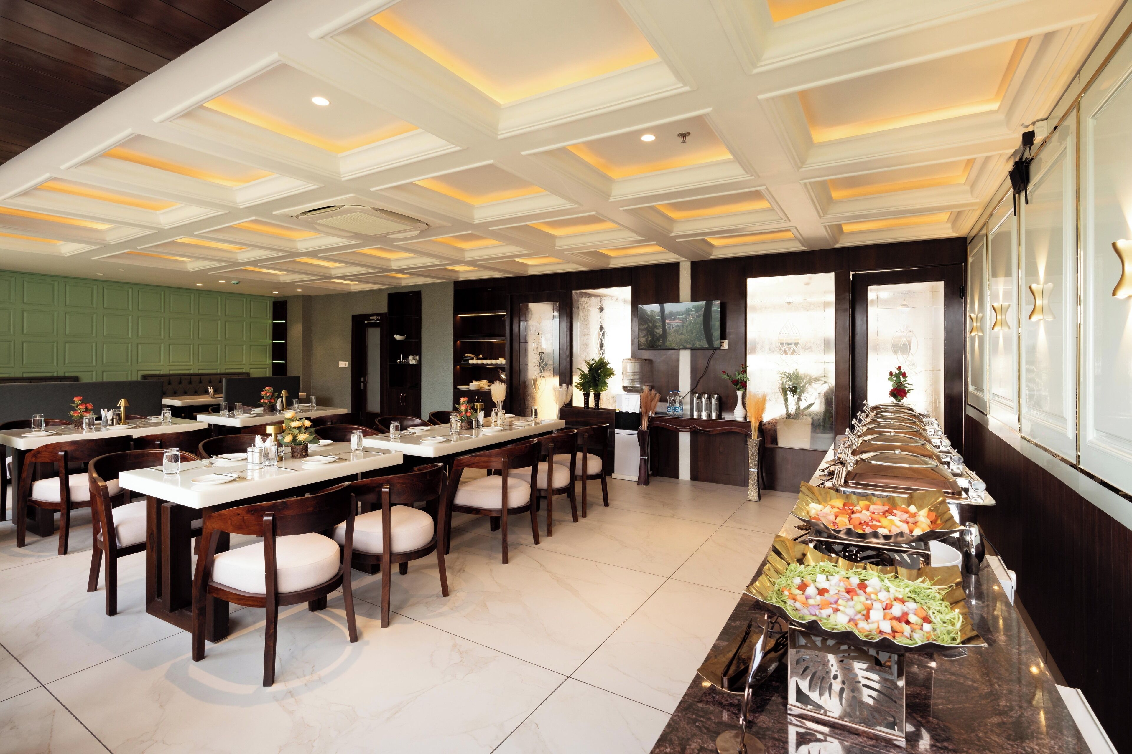 A formal dining area with multiple tables and chairs, a buffet line with various food items, and a television mounted on the wall.