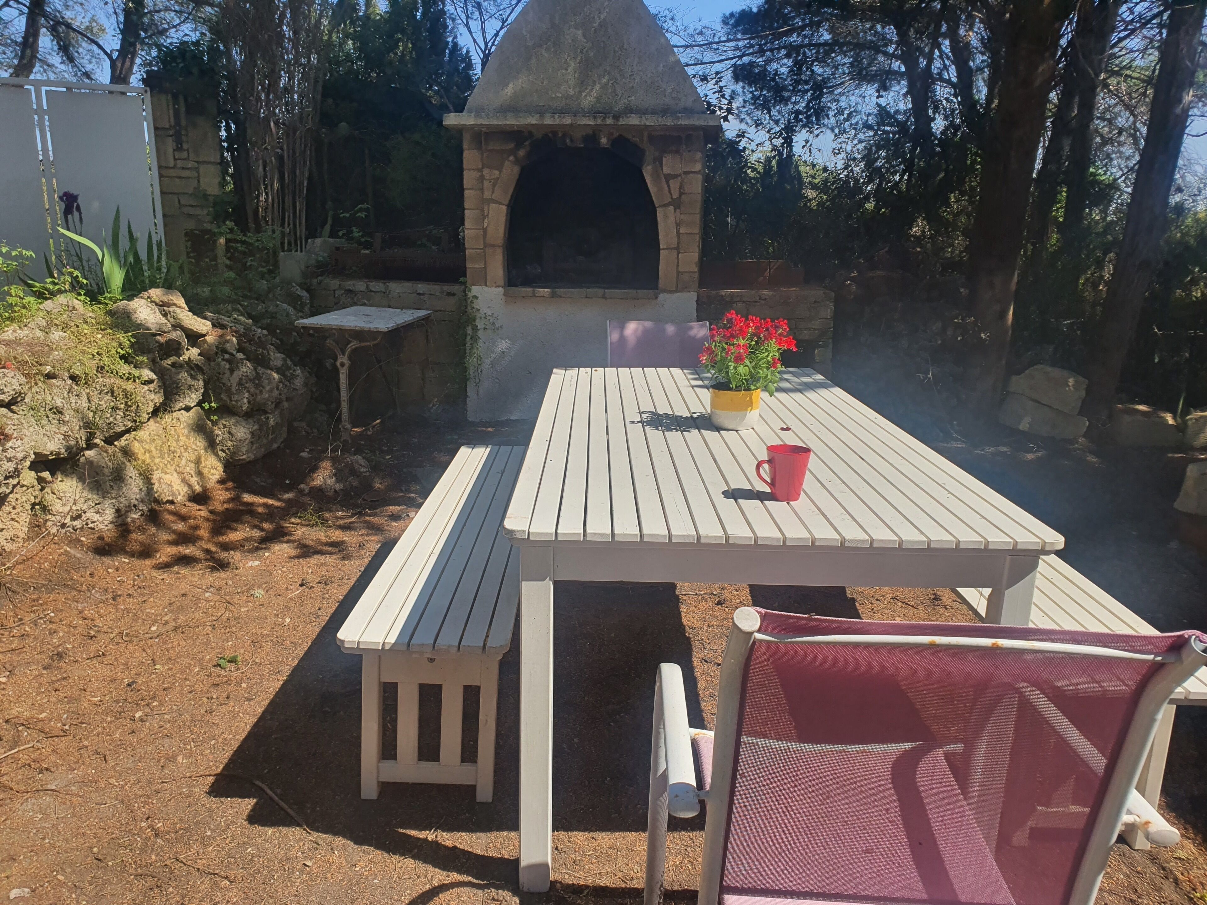 Een terras buiten met een witte tafel en stoelen, een kleine bakoven en een bloempot met rode bloemen.