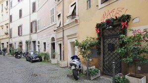 A narrow cobblestone street lined with parked motorcycles and cars, buildings with balconies, and a door decorated with greenery and a red ribbon. - Your nest in Trastevere, the heart of Rome - Your Nest in the Heart of Rome (Roma)