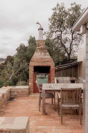 An outdoor brick oven with a chimney, surrounded by a wooden table and chairs, set on a brick patio with trees in the background. - 'Engineers Hut 1823' in Historic Richmond (Richmond)