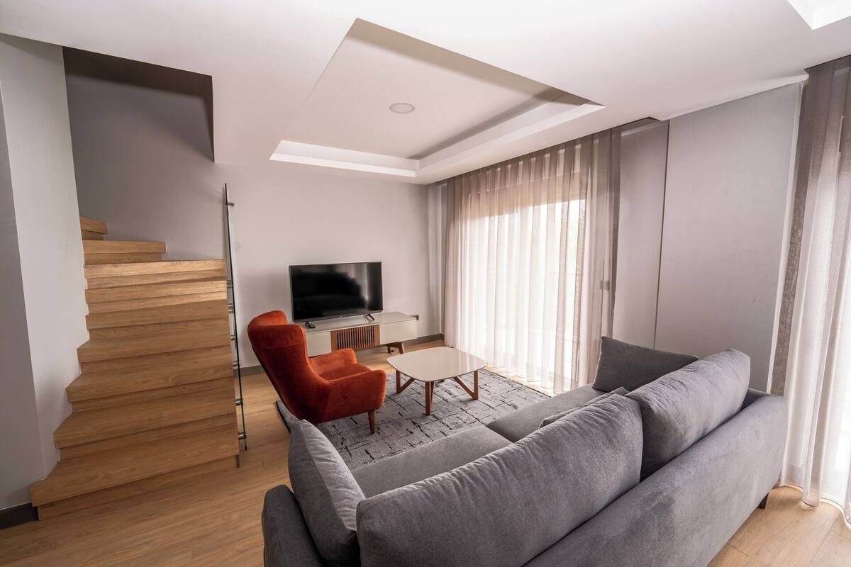 Una sala moderna con un sofá gris, un sillón rojo, un televisor de pantalla plana y una escalera con escalones de madera.
