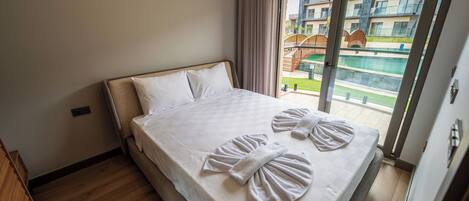 Quarto de hotel com uma cama, janela com vista para um edifício e uma piscina, e um aparelho de ar condicionado na parede.