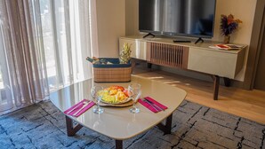 Een moderne hotelkamer met een flatscreen televisie, een eettafel met fruit en glazen, een kleine plant en een houten kast.