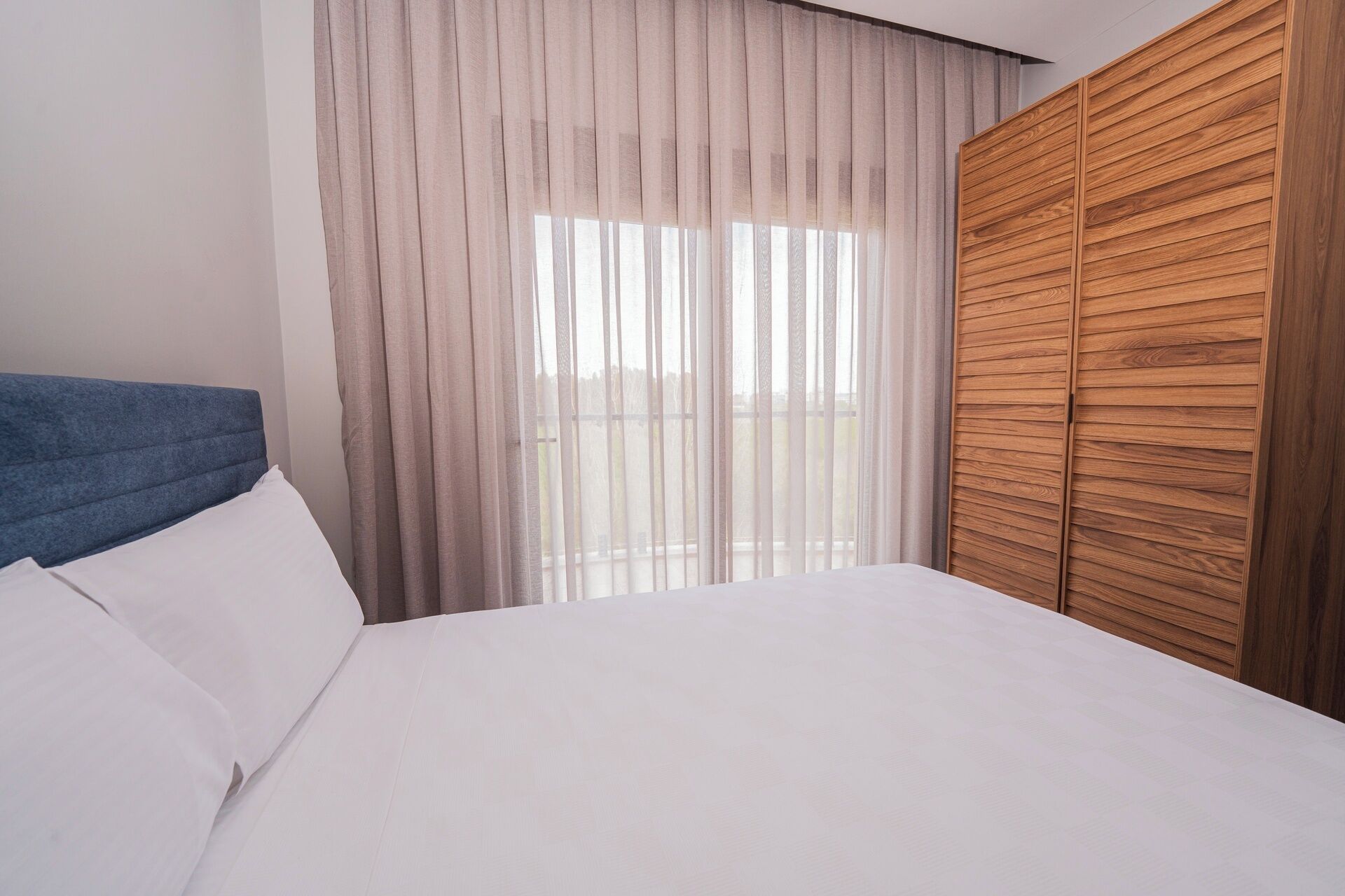 A hotel room with a bed, a wooden wardrobe, and sheer curtains.