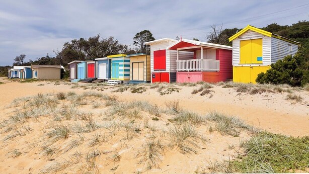 Een rij kleurrijke strandhutten met een zandige voorgrond en schaarse begroeiing.