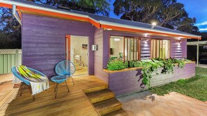 Een modern huis met een paars gevel, een houten terras en een overdekte buiten zithoek met twee stoelen.