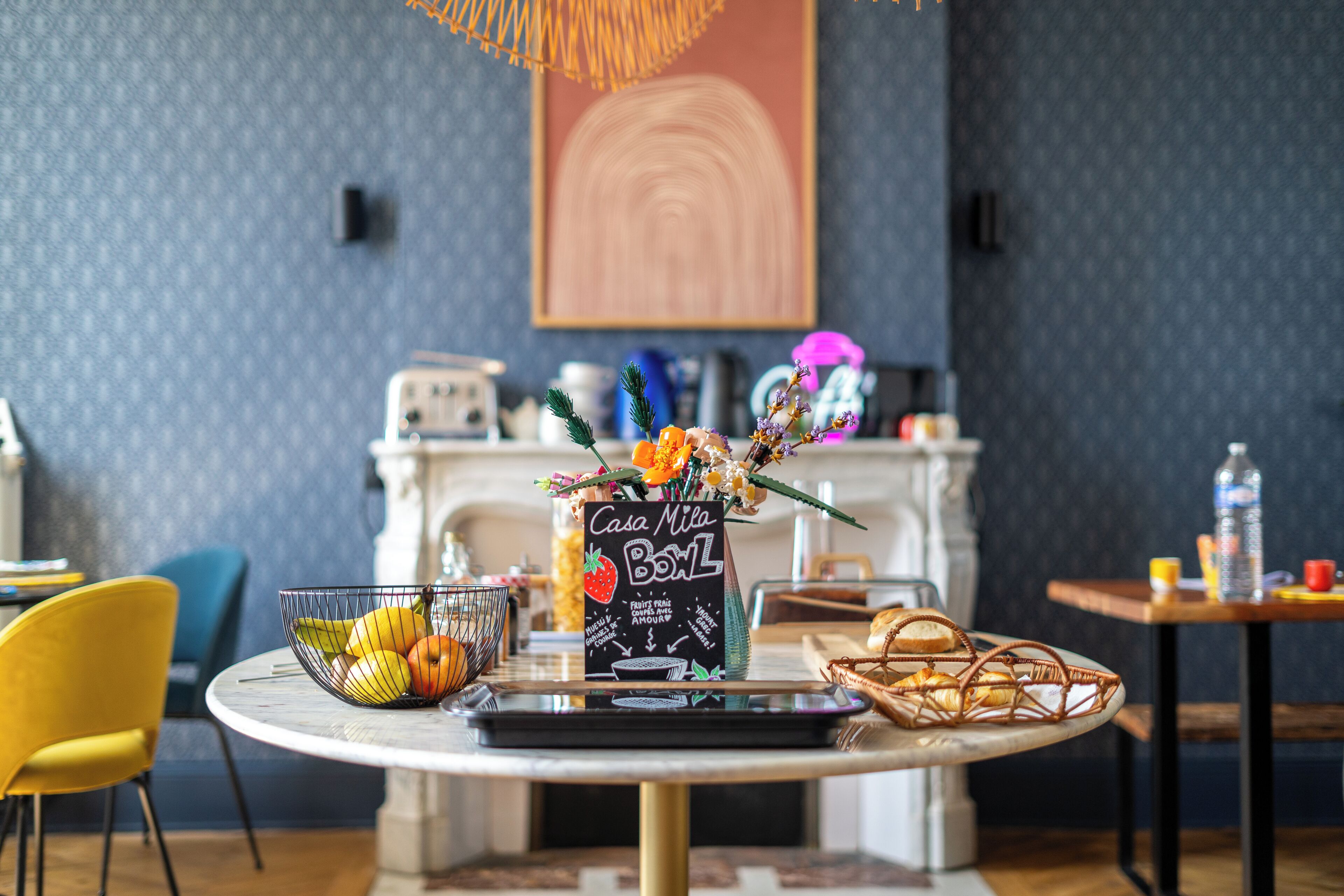 Une table à manger avec un panier de fruits, un panneau décoratif représentant la Casa Mila, et un panier de pain.