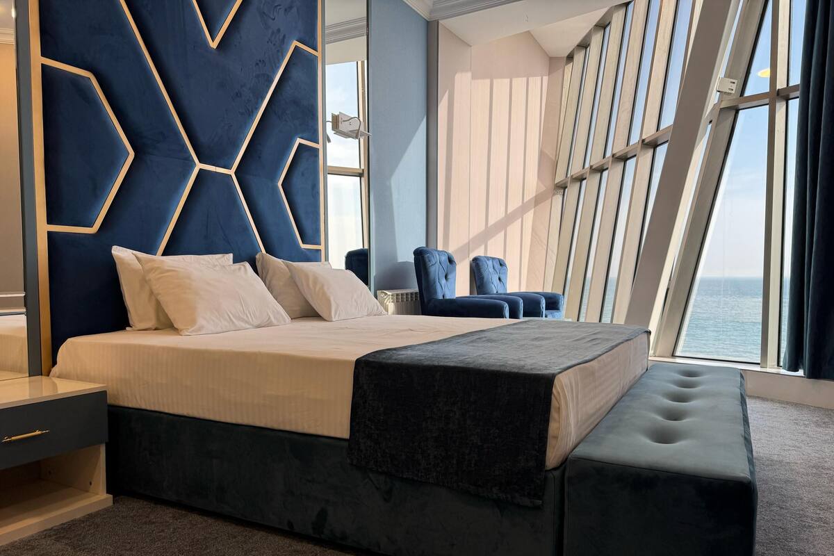A modern bedroom with a large bed, a headboard with geometric patterns, a bench, and a view of the sea through large windows.