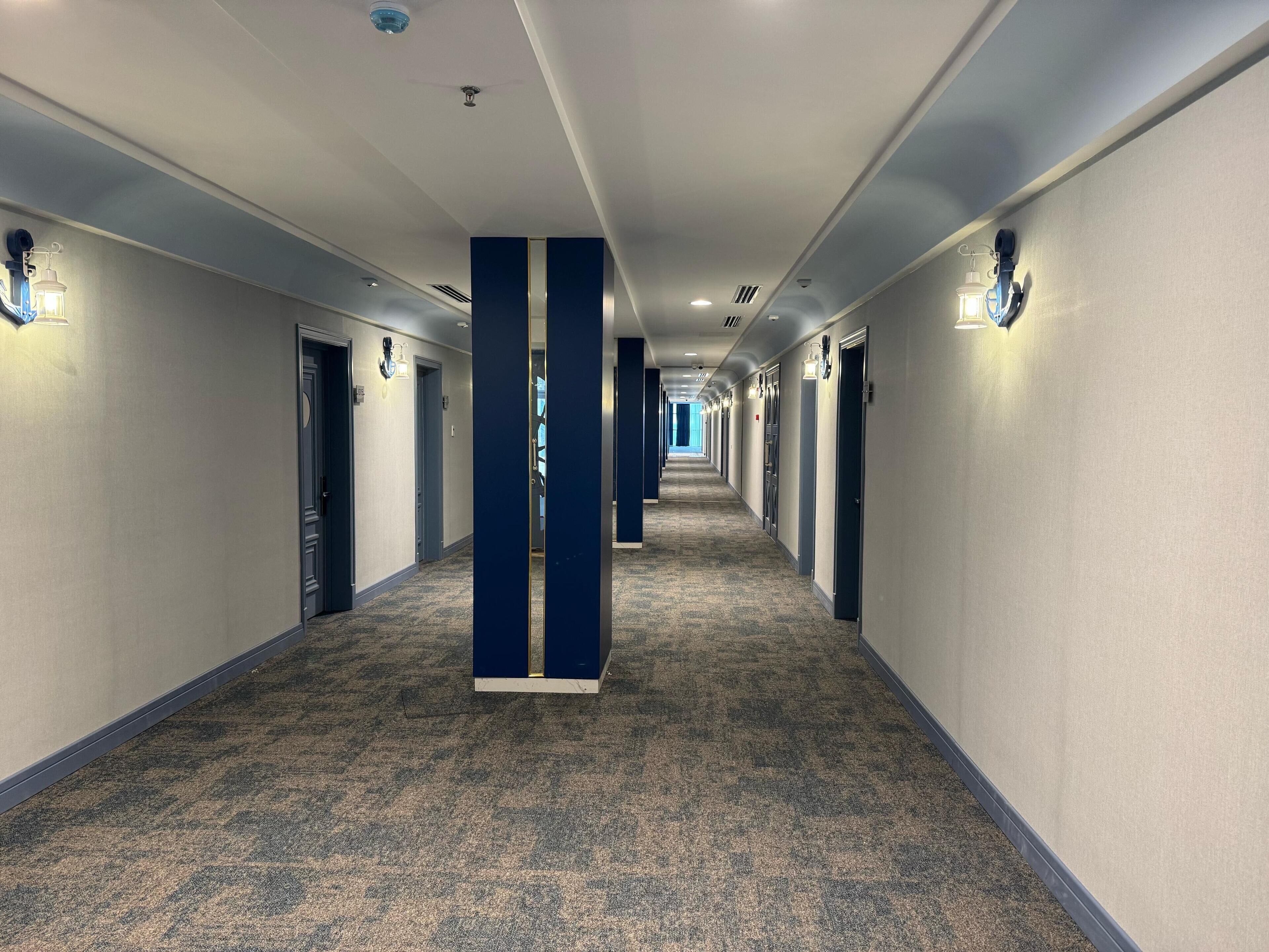A hotel hallway with carpeted floors, blue pillars, and doors with light fixtures.