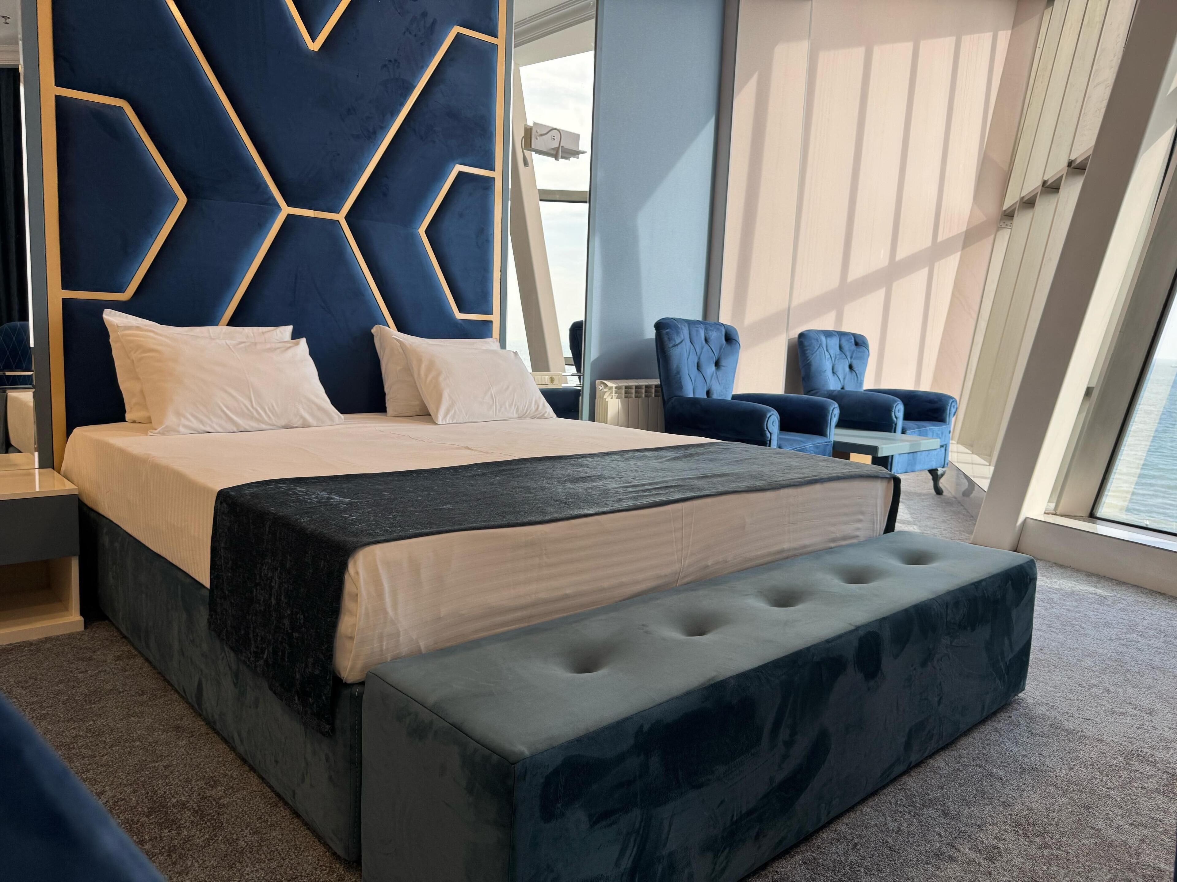 Una habitación de hotel moderna con una cama grande, un cabecero capitonado, dos sillones y vista al mar.